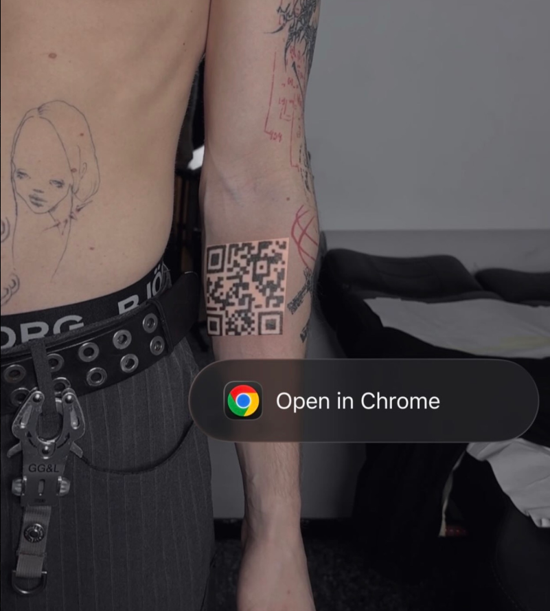 Photograph of the finished QR-code tattoo on skin, freshly inked, matte black modules on a bone-toned background. Every module of the encoded QR is visible in the tattoo.